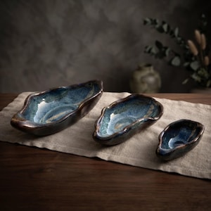 May include: A set of three irregular, boat-shaped ceramic bowls with a glossy finish. The interior features a mottled blend of deep blue, teal, and white glaze, while the exterior is a dark, solid brown. Each bowl has a unique, organic rim.