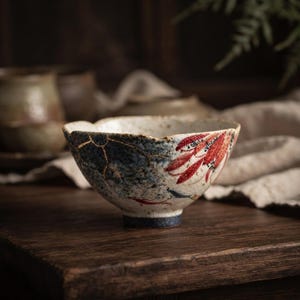 Może przedstawiać: Ceramiczna miska z falistym brzegiem i nakrapianą, złamano-białą podstawą. Zewnętrzna strona ma niebiesko-szare, teksturowane wykończenie ze złotymi liniami w stylu kintsugi oraz wyraźny czerwony wzór kwiatowy namalowany na jednej stronie naczynia.