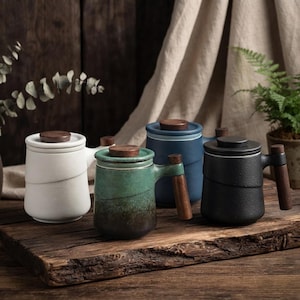 Puede incluir: Un juego de cuatro tazas de té de cerámica con tapa y mango de madera. Las tazas están disponibles en blanco, verde, azul y negro, cada una con un acabado texturizado y forma cilíndrica. Cada taza incluye una tapa a juego con un pomo de madera.