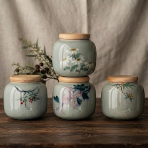 Peut inclure: Quatre bocaux de rangement en céramique vert clair avec une finition émaillée craquelée et des couvercles en bois. Chaque bocal présente des illustrations botaniques peintes à la main, dont des marguerites, des baies rouges et des fleurs.