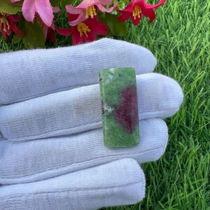 Può includere: Un cabochon di pietra preziosa lucidata di forma rettangolare, caratterizzato da una base verde chiaro screziata con una distinta e irregolare inclusione color magenta scuro centrata sulla superficie, su uno sfondo di erba verde e fiori rosa.