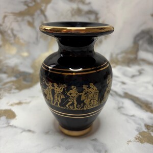May include: A black ceramic vase with gold trim and detailing. The vase features a classical design with gold figures and accents. The top and base of the vase are trimmed in gold.