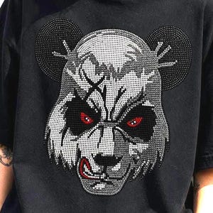 Puede incluir: Una camiseta negra de manga corta con un gráfico grande y detallado de una cara de panda en la parte delantera. El diseño del panda está compuesto por pequeños diamantes de imitación brillantes en tonos plateados y grises, con ojos y lengua rojos.