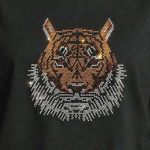 Könnte beinhalten: Ein schwarzes T-Shirt mit kurzen Ärmeln und einem zentralen Motiv eines Tigerkopfes, der aus kleinen, reflektierenden Strasssteinen besteht. Das Tigerdesign enthält braune, orangefarbene und weiße Kristalle in einem geometrischen Muster.