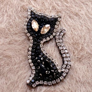May include: A black cat brooch with a rhinestone border and accents. The cat's body is filled with black rhinestones, and the eyes are two amber-coloured stones. The brooch is set against a neutral, textured background.