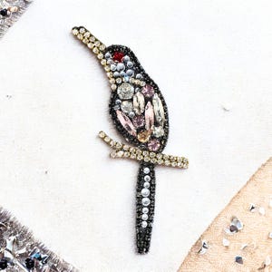 Puede incluir: Broche decorativo de pájaro con cuerpo negro adornado con cristales transparentes, rosas y rojos. El pájaro está posado en una rama dorada, con pedrería dorada en la cabeza y la rama.