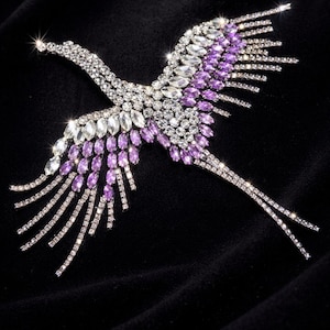 May include: A glittering bird brooch, crafted from clear and purple crystals, set against a black backdrop. The bird is depicted in mid-flight, with extended wings and a slender neck. The crystals shimmer, producing a radiant display.
