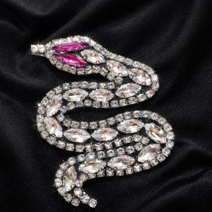 Puede incluir: Un broche de serpiente brillante, adornado con pedrería. La cabeza de la serpiente presenta gemas rosas, mientras que el cuerpo está adornado con pedrería transparente en forma ovalada. El broche se encuentra sobre un fondo negro.