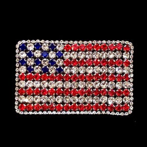 Op de afbeelding: Een rechthoekige broche met het ontwerp van de Amerikaanse vlag. De vlag is samengesteld uit rode, witte en blauwe strass-steentjes, met een rand van heldere strass-steentjes. De achtergrond is zwart.