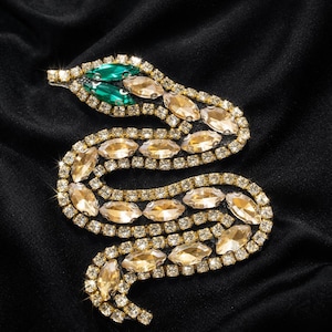 Peut inclure: Un motif de serpent orné de pierres précieuses, avec un cadre doré et des pierres transparentes et ambrées. La tête du serpent est ornée de pierres vertes, sur fond noir.