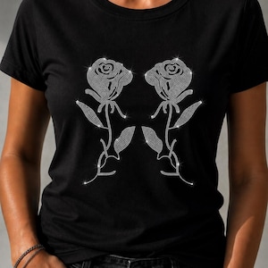 May include: A black short-sleeved t-shirt featuring a graphic design of two mirrored roses created with small, reflective silver-toned rhinestones. The floral pattern is centered on the chest, showcasing detailed petals, stems, and leaves.