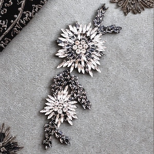 May include: Two floral embellishments with clear and dark grey crystals. The larger flower has a central cluster of clear crystals. Dark grey crystal leaves and stems connect the flowers. The background is a light grey fabric.
