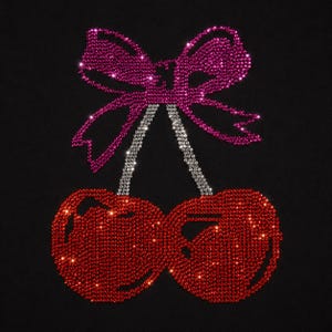 May include: A rhinestone applique on a black background featuring two red cherries with silver stems and a large pink bow tied at the top. The design is composed of small, reflective crystals that create a sparkling effect across the entire surface.