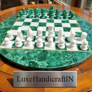 Puede incluir: Un juego de ajedrez redondo con un tablero verde malaquita y blanco. Las piezas son blancas y verdes. El tablero está sobre una mesa de madera. La placa plateada muestra el texto "LuxeHandicraftIN".
