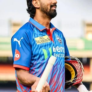May include: A man in a blue and red cricket jersey with the text "All Seasons" and "Hero Encor". He is holding a cricket bat and helmet. The jersey has a blue and red pattern. He is wearing a black headband.