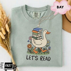 Puede incluir: Camiseta verde claro con un pato de dibujos animados que lleva libros, rodeado de flores. El texto "LET'S READ" está debajo del pato. Un collar plateado con un colgante de estrella está en la camiseta.