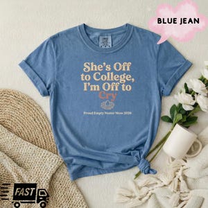 May include: A blue short-sleeve t-shirt with the text "She's Off to College, I'm Off to Cry" in cream and orange lettering, featuring a small nest graphic and the text "Proud Empty Nester Mom 2026" printed on the front center.