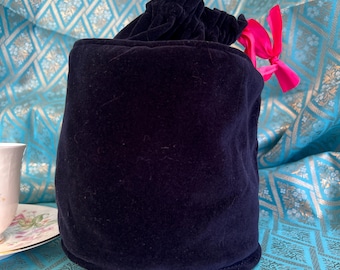 May include: A cylindrical dark navy blue velvet drawstring pouch featuring a gathered top closure secured with a bright pink ribbon. The pouch has a flat circular base and a soft, plush texture, standing upright as a container for small items.