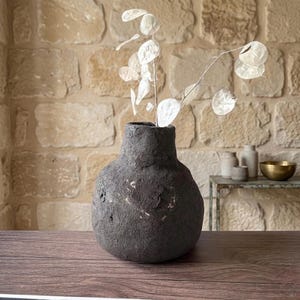 May include: A dark gray, textured vase with a round body and narrow neck, holding delicate, white, circular dried flowers. The vase sits on a wooden surface against a stone wall background. Decorative home decor.