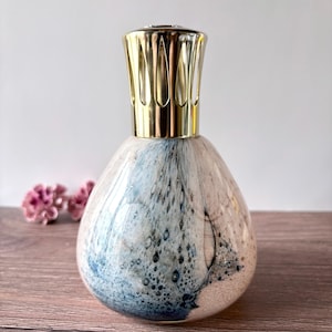 Op de afbeelding: Een druppelvormige geurlamp van glas met een gevlekt patroon in wit, zachtroze en diepblauwe tinten. De houder is afgewerkt met een gepolijste goudkleurige metalen kroonkap met verticale uitsparingen voor een decoratieve uitstraling.