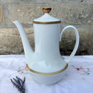 May include: A white porcelain coffee pot featuring a tall, cylindrical body with a curved handle and a long, slender spout. The lid is topped with a small, metallic gold-toned knob, and the rim and base are decorated with a textured gold band.
