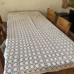 May include: A rectangular wooden table with a white, open-weave lace tablecloth. The table has a rustic appearance with visible wood grain. Two woven-back chairs are visible. The room has a neutral color palette.