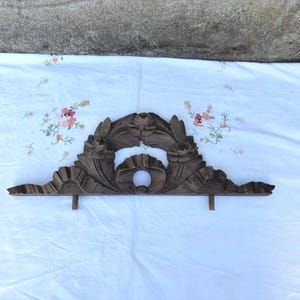 Può includere: Un frontone architettonico in legno marrone scuro con intricati motivi floreali intagliati a mano, tra cui rose centrali e motivi a foglia. Il pezzo ha una forma arcuata simmetrica con due piccoli tasselli in legno rivolti verso il basso.