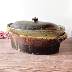 May include: An oval ceramic casserole dish with a dark brown and black glaze, featuring a matching lid with a central knob and two side handles. The exterior displays a textured, earthy finish with a lighter yellowish-green band near the rim.