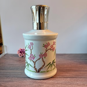 May include: A white ceramic fragrance lamp with a cylindrical body featuring a painted design of brown branches, pink blossoms, and green bamboo leaves. The top of the lamp is fitted with a polished silver-colored metal burner cap.