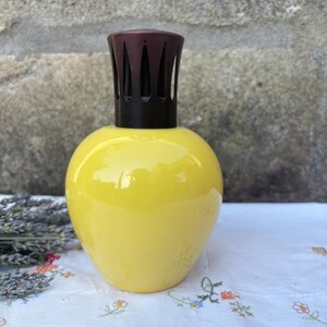 May include: A bright yellow, rounded glass fragrance lamp with a smooth, glossy finish. The top features a dark brown, crown-shaped metal cap with vertical cutouts, designed to sit securely on the neck of the bulbous glass vessel.