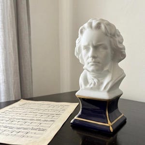 Peut inclure: Buste en porcelaine blanche d'un homme aux cheveux bouclés, posé sur une base bleu marine avec des bordures dorées. Le buste est placé sur une surface en bois sombre à côté d'une partition de musique vieillie. Une œuvre d'art classique.