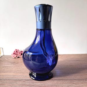 Op de afbeelding: Een druppelvormige geurlamp van kobaltblauw glas met een brede basis en een smalle hals. De bovenkant is voorzien van een matte donkerblauwe metalen kroon met verticale uitsparingen. Een dikke, donkerblauwe stoffen lont loopt in de glazen houder.