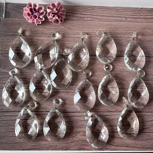 Peut inclure: Un ensemble de treize cristaux en verre transparents, facettés et en forme de goutte, chacun doté d'une petite boucle métallique au sommet pour la suspension. Deux petites fleurs en tissu plissé rose poudré sont placées en haut à gauche.