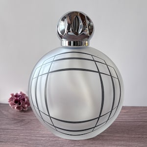 Peut inclure: Un flacon diffuseur de parfum sphérique en verre dépoli avec un motif de grille gris foncé sur le devant. Le flacon est surmonté d'un bouchon en métal argenté poli doté d'un design décoratif à facettes multiples et ajouré.