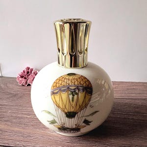 Può includere: Una lampada profumata sferica in ceramica bianca con un'illustrazione dipinta a mano di una mongolfiera gialla e marrone sul davanti. La lampada è sormontata da un tappo a corona in metallo color oro lucido con aperture verticali.