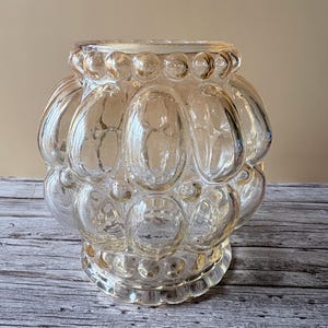 Peut inclure: Un vase en verre transparent avec un motif de bulles. Le vase a une forme arrondie avec une base large et un sommet légèrement plus étroit. Le verre a une légère teinte ambrée, et les bulles sont espacées uniformément.