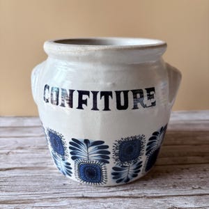 Peut inclure: Un pot en céramique blanc avec le mot "CONFITURE" en lettres bleu foncé. Le pot présente un motif floral bleu répétitif autour de la base et possède deux petites poignées. Le pot est probablement utilisé pour conserver des confitures.