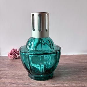 Op de afbeelding: Een blauwgroene glazen geurlamp met een afgerond, geribbeld lichaam en een brede basis. De bovenkant is voorzien van een cilindrische zilverkleurige metalen branderkap met verticale uitsparingen. Een witte katoenen lont is binnenin zichtbaar.