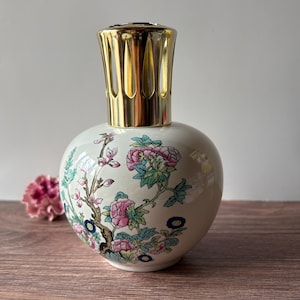 May include: A white ceramic fragrance lamp with a rounded body featuring a colorful floral pattern of pink peonies and green leaves. The lamp is topped with a gold-colored metal crown-shaped burner cap with vertical cutouts for ventilation.
