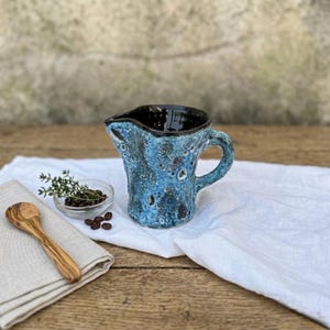 May include: A blue ceramic pitcher with a textured surface and a black interior. The pitcher has a handle and a spout. A small wooden spoon, a small glass bowl with herbs, and coffee beans are on a white cloth.