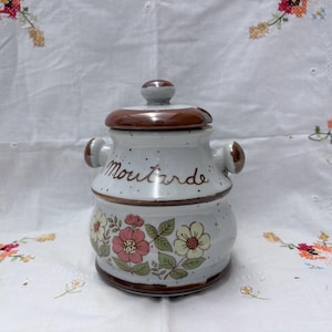 Op de afbeelding: Een witte keramische mosterdpot met een bruin deksel en handvatten. De pot is versierd met roze en witte bloemen en groene bladeren. Het woord "Moutarde" staat op de pot.