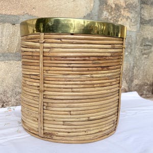 Può includere: Un cestino o contenitore cilindrico realizzato in canne di rattan intrecciate color marrone chiaro. Il contenitore presenta una fascia liscia in ottone lucido color oro che circonda il bordo superiore, creando un contrasto con il materiale naturale.