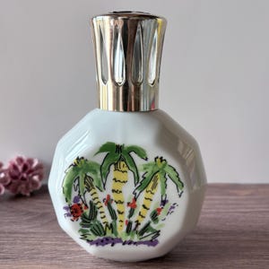 Op de afbeelding: Een witte keramische geurlamp met een achthoekig lichaam voorzien van een handgeschilderd ontwerp van gele en groene palmbomen met kleine rode accenten. De lamp is afgewerkt met een geventileerde dop van gepolijst zilverkleurig metaal.
