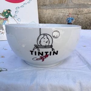 Peut inclure: Un bol en céramique blanche orné d'un dessin au trait noir représentant le personnage Tintin portant un casque spatial, avec le mot TINTIN imprimé en lettres noires grasses en dessous. Un petit motif de fusée rouge et blanc est situé sous le texte.