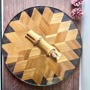 May include: A circular tray featuring a geometric star pattern composed of light tan and dark brown bamboo strips. Centered on the tray is a cylindrical bamboo handle with two distinct nodes, showcasing the natural texture and light tan colour.