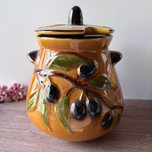 Può includere: Un barattolo in ceramica smaltata con coperchio, caratterizzato da una base color ambra caldo decorata con un motivo in rilievo di olive nere e foglie verdi su un ramo marrone. Il barattolo ha due piccole maniglie laterali e un pomello centrale.
