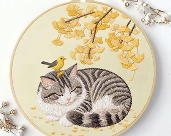 May include: A circular embroidery hoop containing a cream-coloured fabric with a hand-stitched design of a sleeping grey and white tabby cat. A small yellow bird rests on the cat's head, positioned beneath embroidered yellow ginkgo leaves.