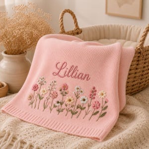 May include: A soft pink knit baby blanket featuring the embroidered name Lillian in cursive script above a colourful row of stitched wildflowers in shades of white, yellow, and pink with green stems and leaves, finished with a ribbed edge.