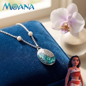 Peut inclure: Un collier argenté avec un pendentif turquoise et argenté, posé sur un coussin de velours bleu. Le collier a une chaîne avec deux perles blanches. Le mot "Moana" est visible en haut à gauche.