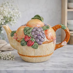 May include: A ceramic teapot with a fruit design. The teapot is beige with a wood-grain texture and is decorated with peaches, grapes, and strawberries. The handle and spout are a light brown colour. The teapot is sitting on a marble surface.